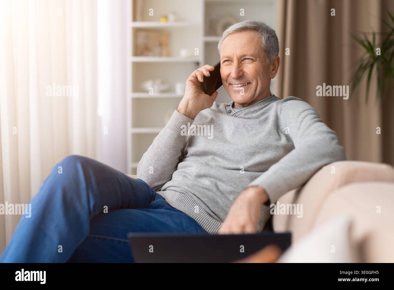 Happy senior man sitting comfortably on sofa at home talking on smartphone Stock Photo