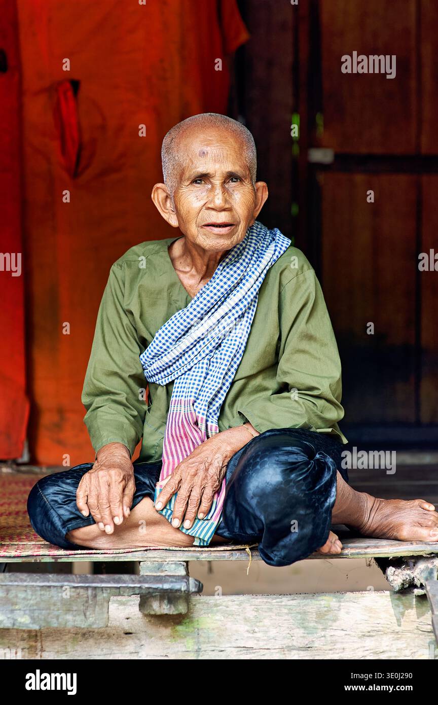 An elderly man with a shaved head sits on a mat. He wears traditional clothing and poses for a portrait in his home. Stock Photo