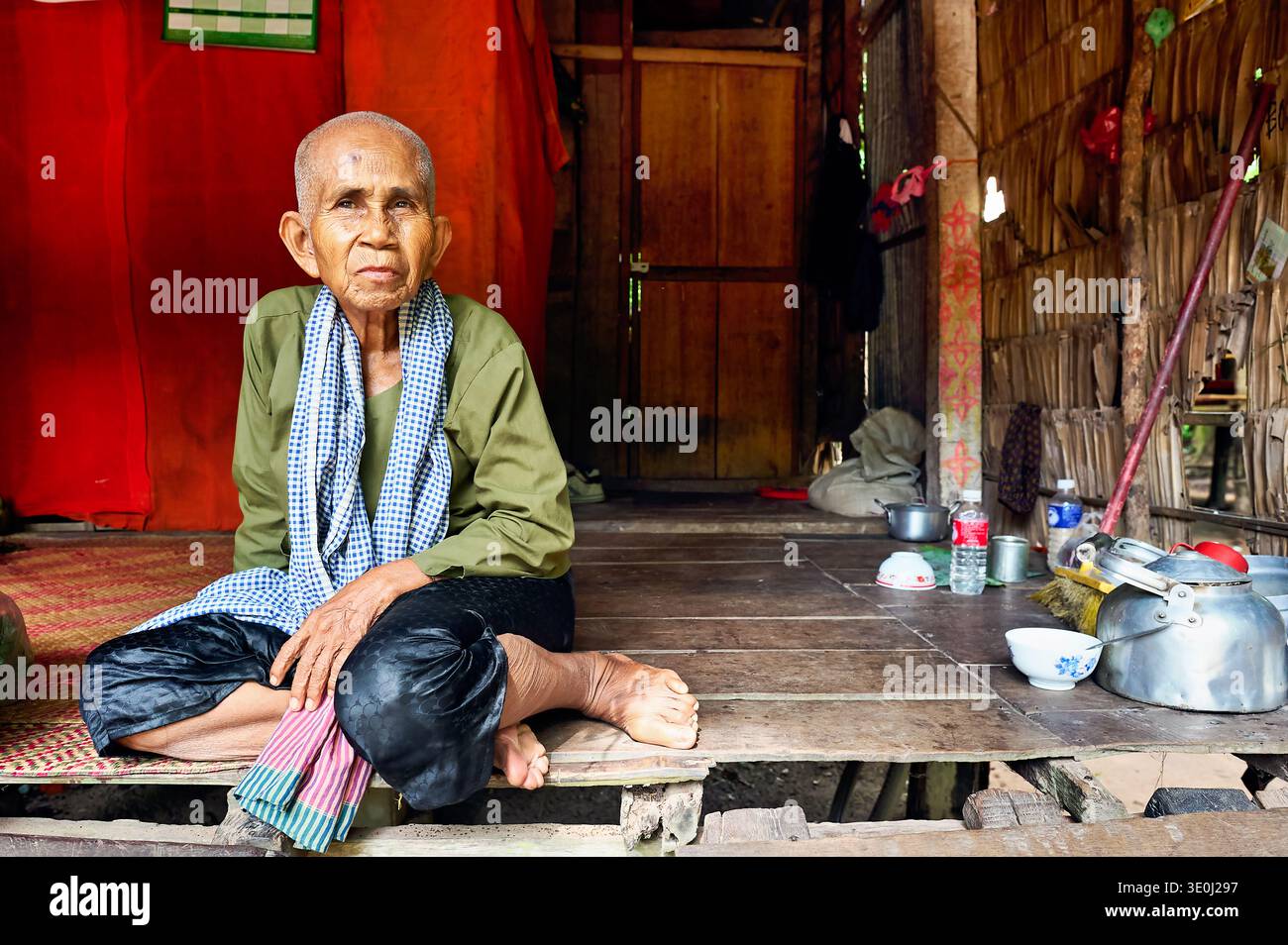 An elderly Cambodian woman in traditional attire sits on a mat inside her home. She relaxes and enjoys her surroundings during the day. Stock Photo