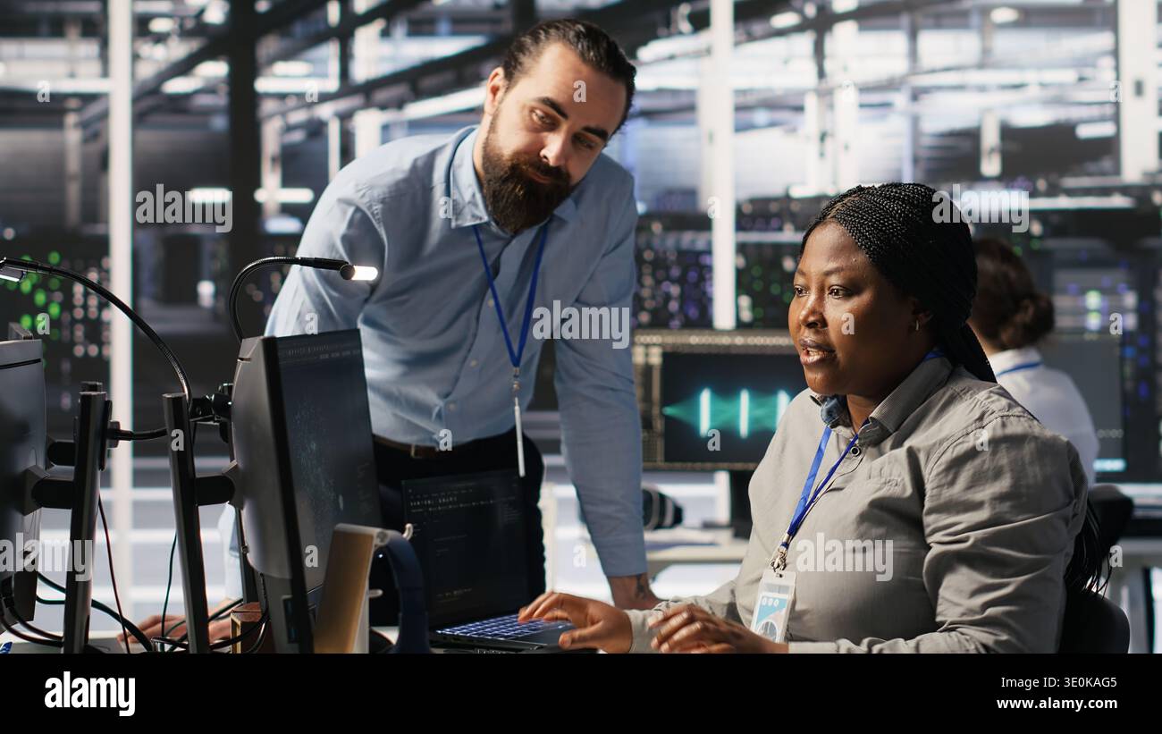 Server room manager providing feedback to employee managing AI training datasets. Team leader talking with technician in data center supervising system operations supporting neural networks, camera A Stock Photo