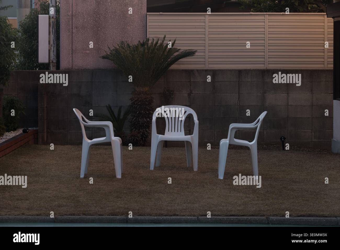 Three white plastic chairs arranged on a dry lawn in a quiet residential courtyard Stock Photo