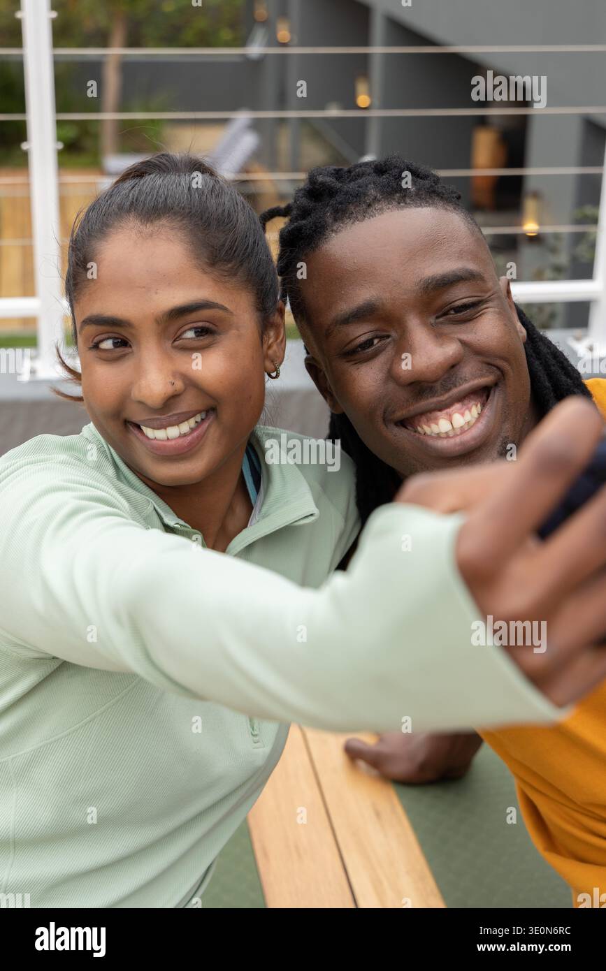 Diverse couple posing and leaning close on rooftop patio, holding smartphone on wooden bench Stock Photo