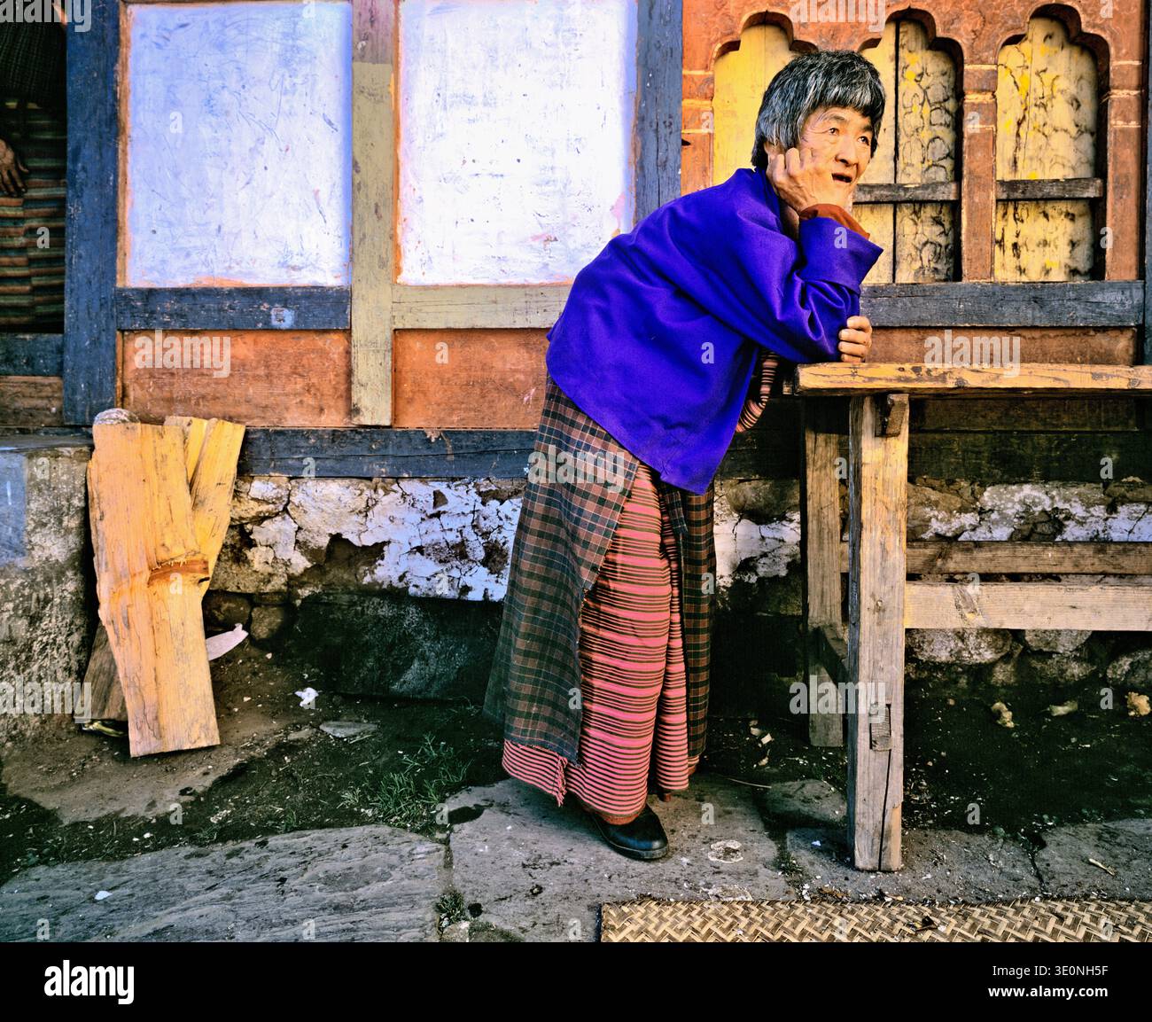 An elderly woman in traditional attire leans on a wooden table and watches her village with a gentle expression during the day. Stock Photo