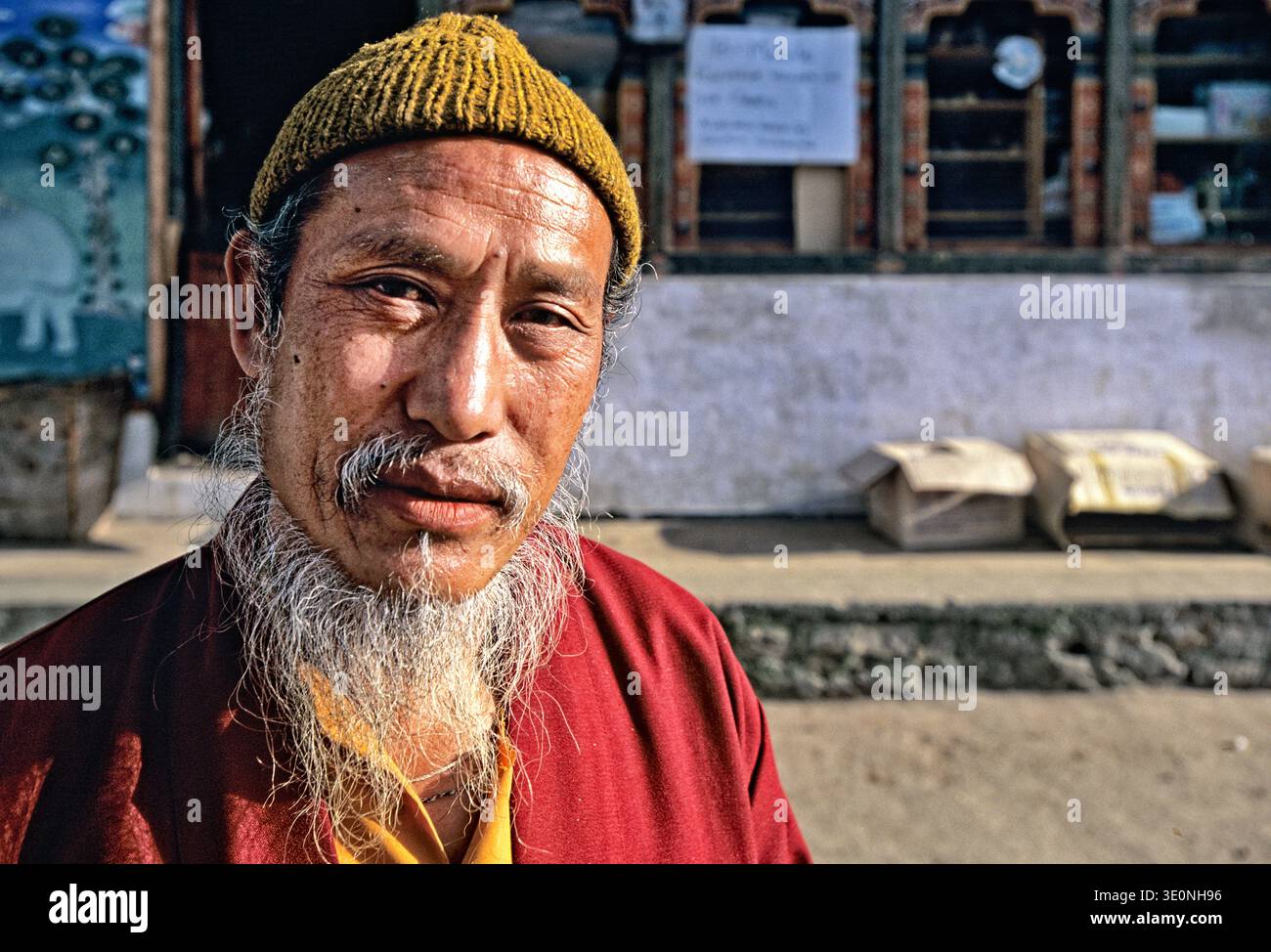 An elderly man in traditional dress stands on a street in Bhutan. He has a long white beard and wears a yellow beanie. Stock Photo