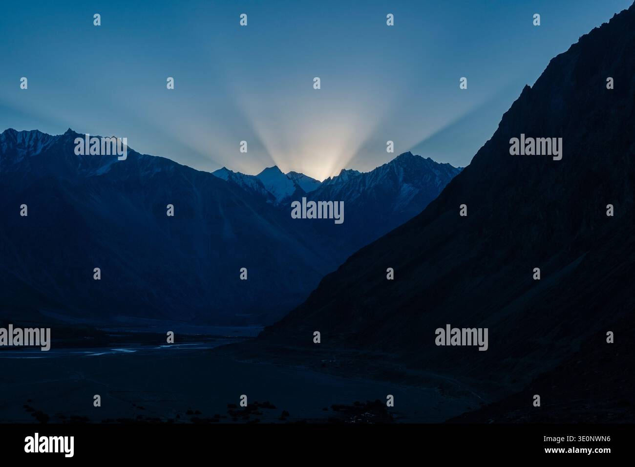 Bright sunbeams radiate from behind the dark, jagged silhouettes of the Karakoram mountains during sunrise in Diskit. Stock Photo