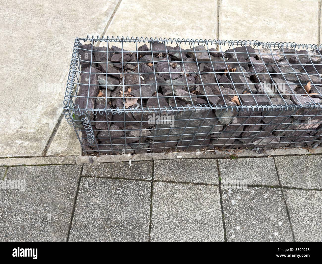 Close up view of a metal wire mesh gabion basket filled with dark stones resting against concrete paving slabs - Smartphone Captured Stock Image