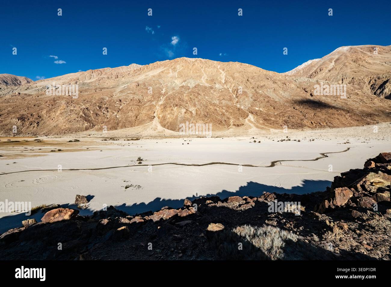 A narrow stream winds through the vast, arid Nubra Valley floor towards the steep, rocky slopes of the Karakoram Range in the distance. Stock Photo