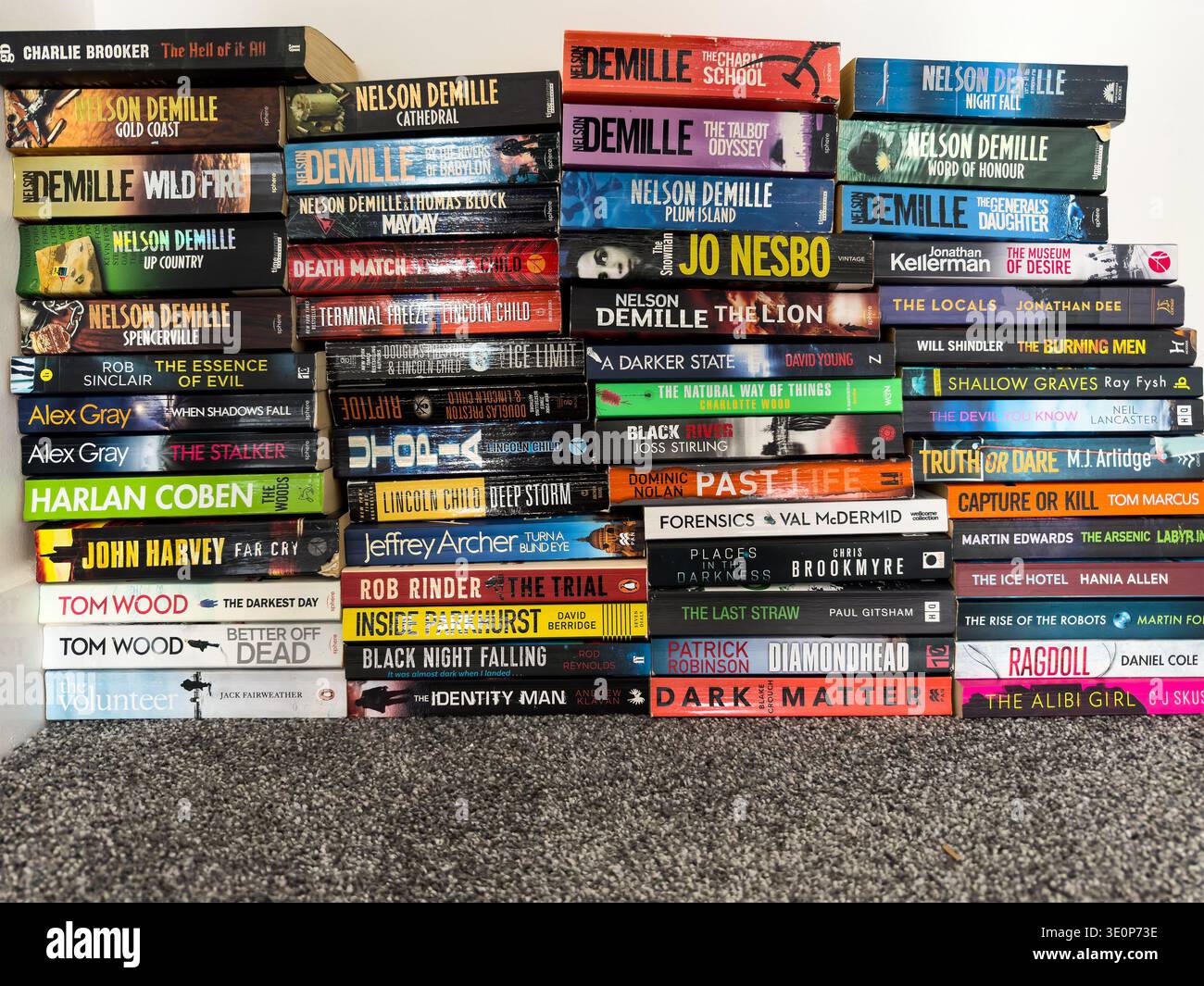 Large collection of thriller and crime paperback books stacked vertically on a carpeted surface with a white wall above - Smartphone Captured Stock Image