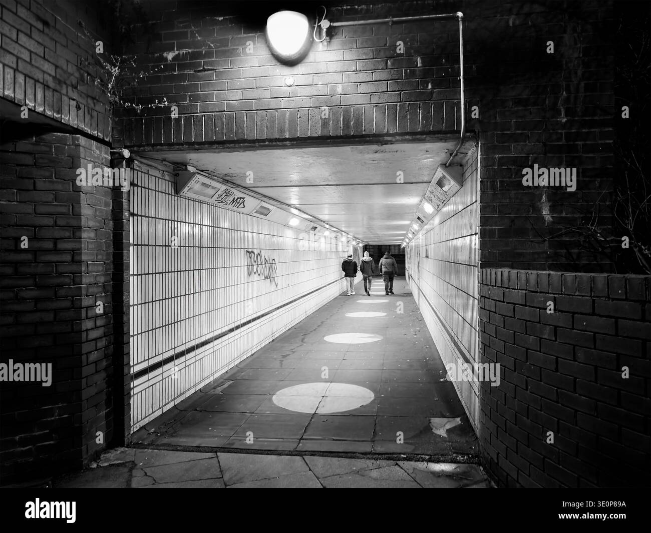 Three figures walk through a dimly lit pedestrian underpass at night with exposed brickwork and tiled walls in Coventry uk - Smartphone Captured Stock Image