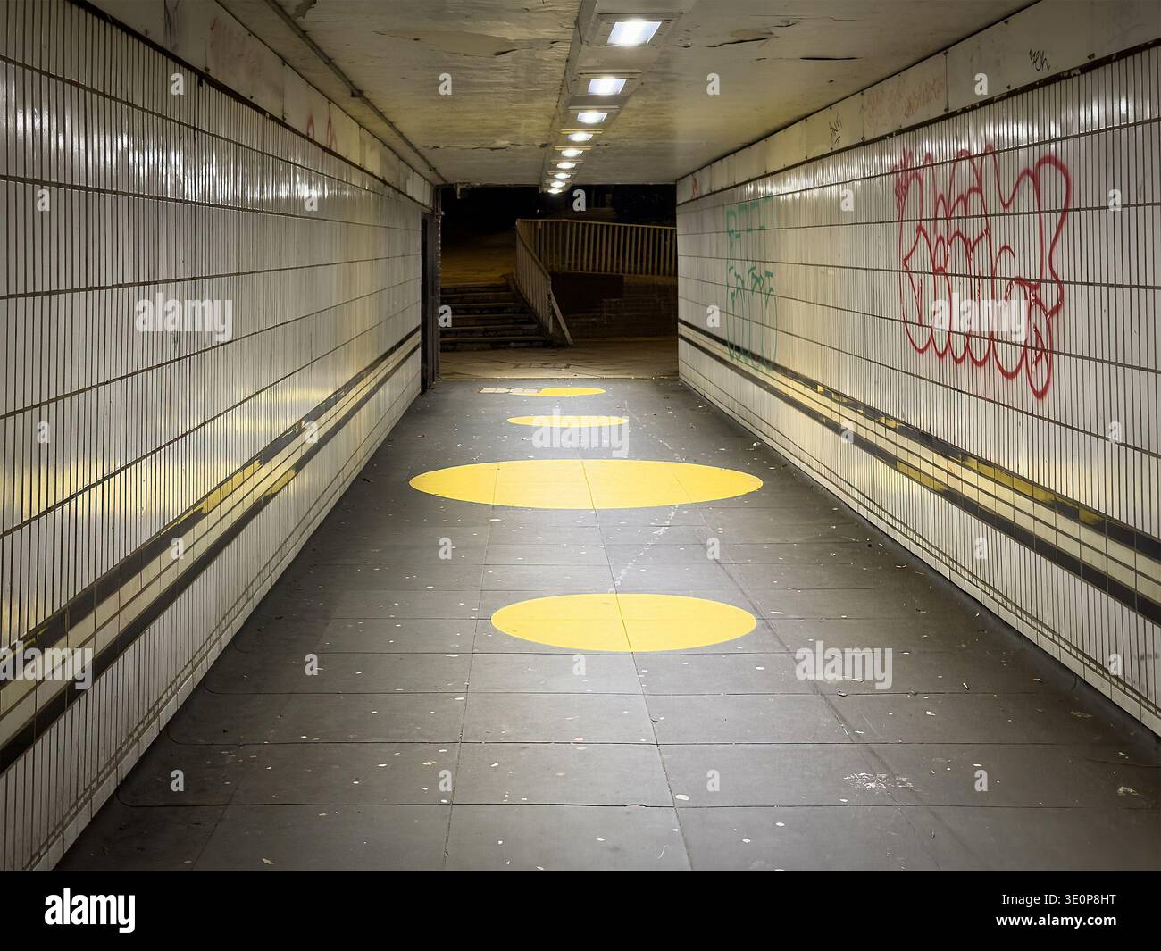 Dimly lit pedestrian underpass illuminated by overhead lights with yellow circular floor markings and graffiti on the walls  in Coventry uk - Smartphone Captured Stock Image