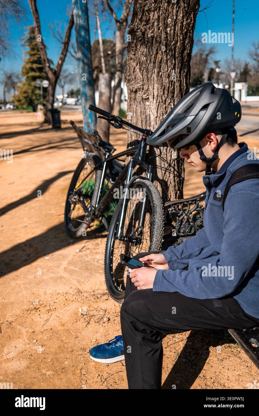 Young man wearing helmet sitting on a bench in a park, using his cell phone with his mountain bike nearby Stock Photo