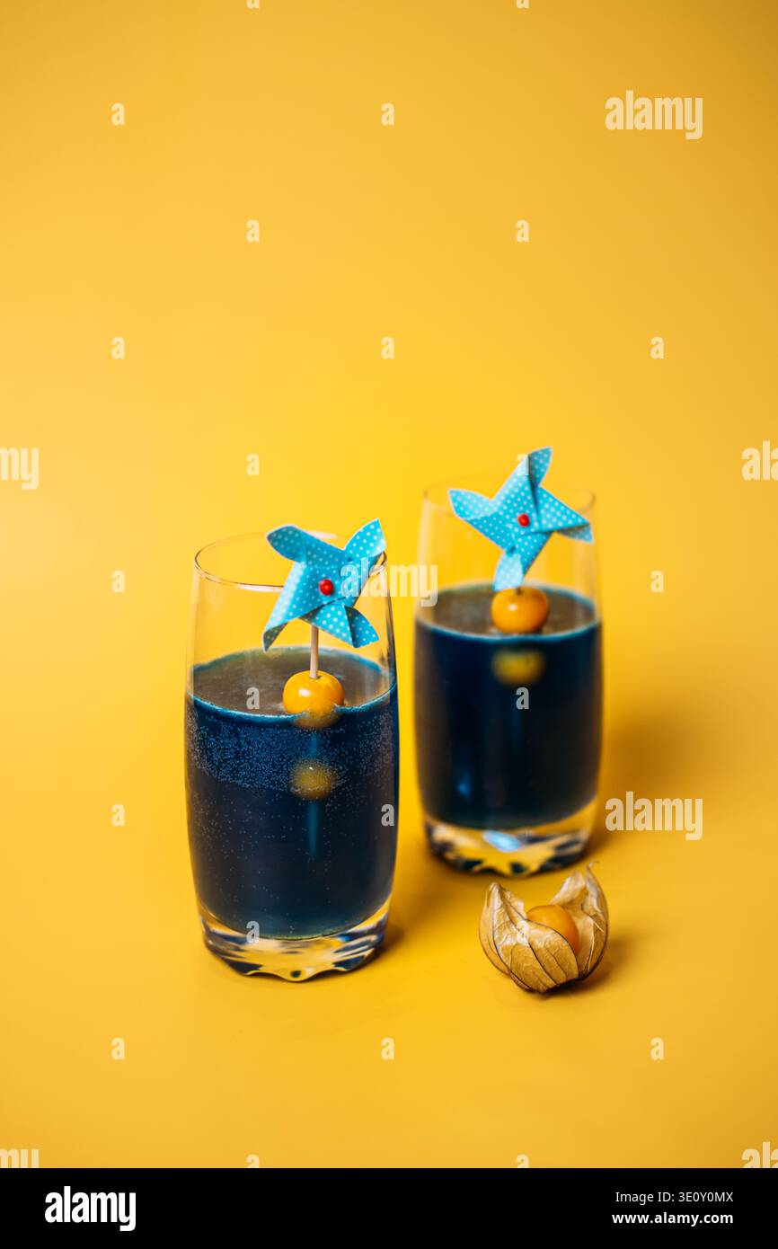 Two glasses of deep blue liquid decorated with pinwheels and physalis sit on yellow background. physalis cocktail garnish, cape gooseberry drinks Stock Photo