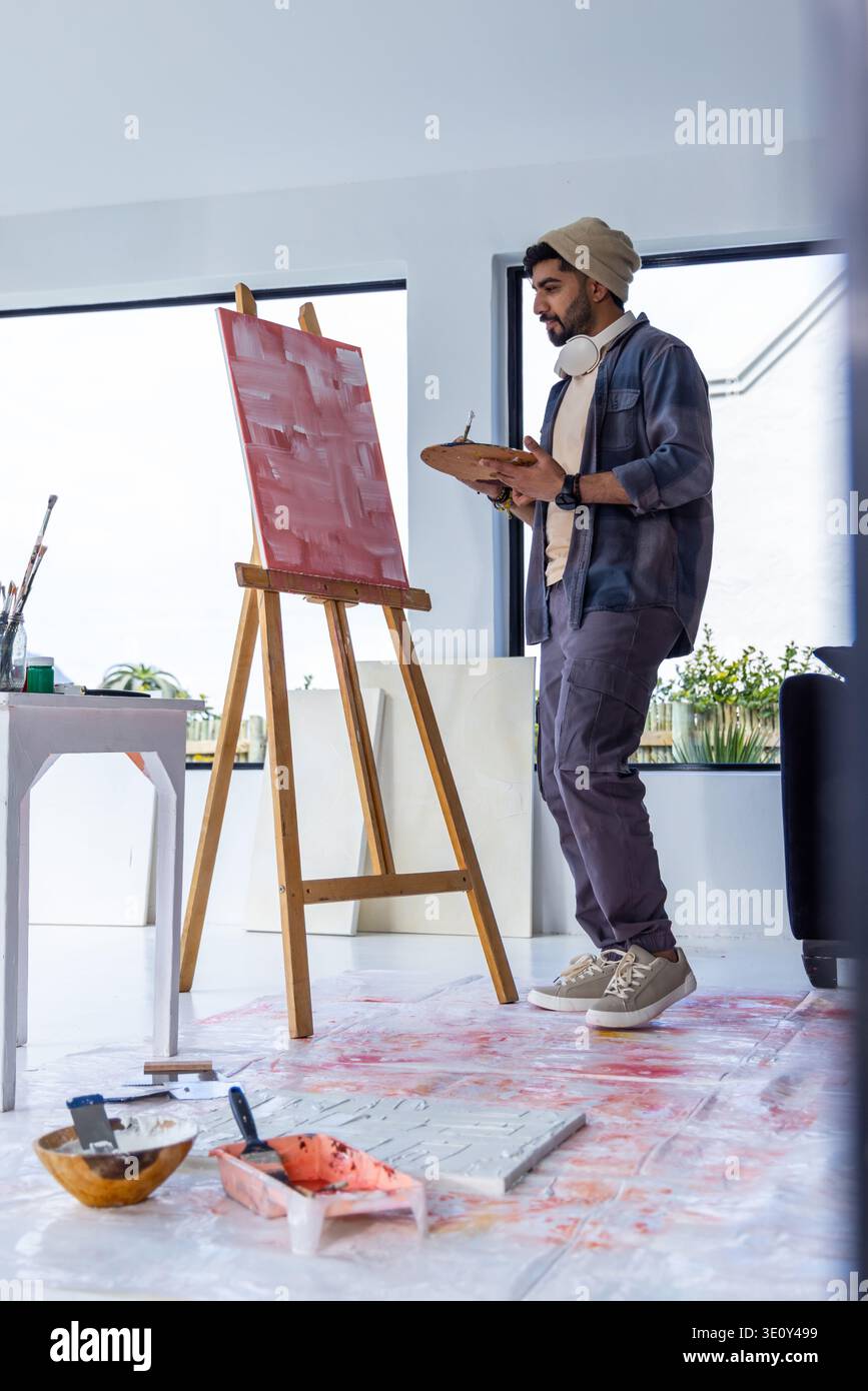 Male artist standing in bright art studio working at wooden easel with paint palette, copy space Stock Photo