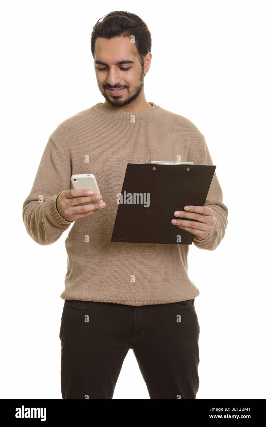 Young happy Indian man holding clipboard while using mobile phone Stock Photo