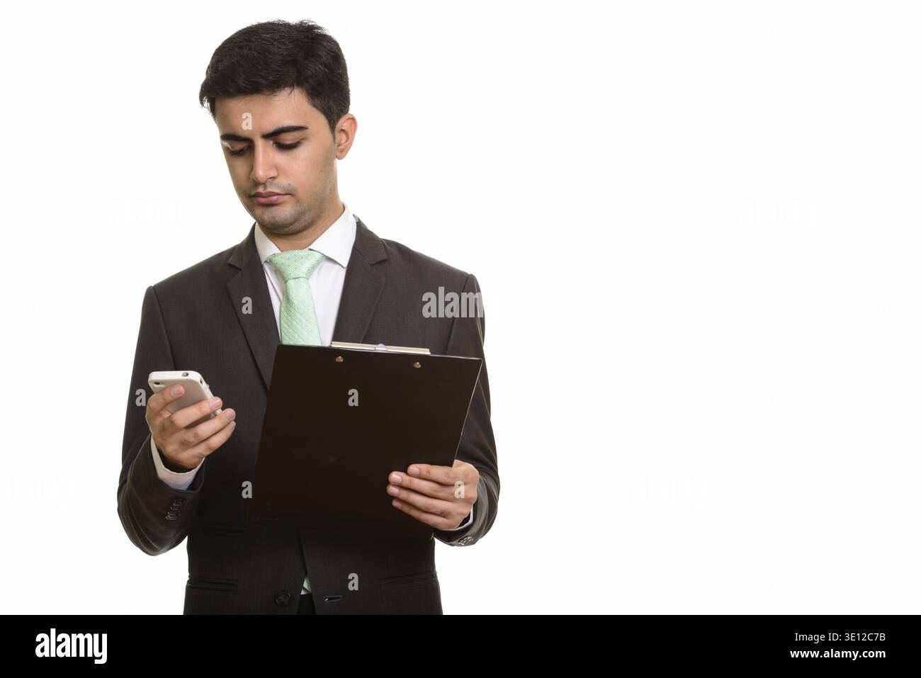Young handsome Persian businessman holding clipboard while using mobile phone Stock Photo