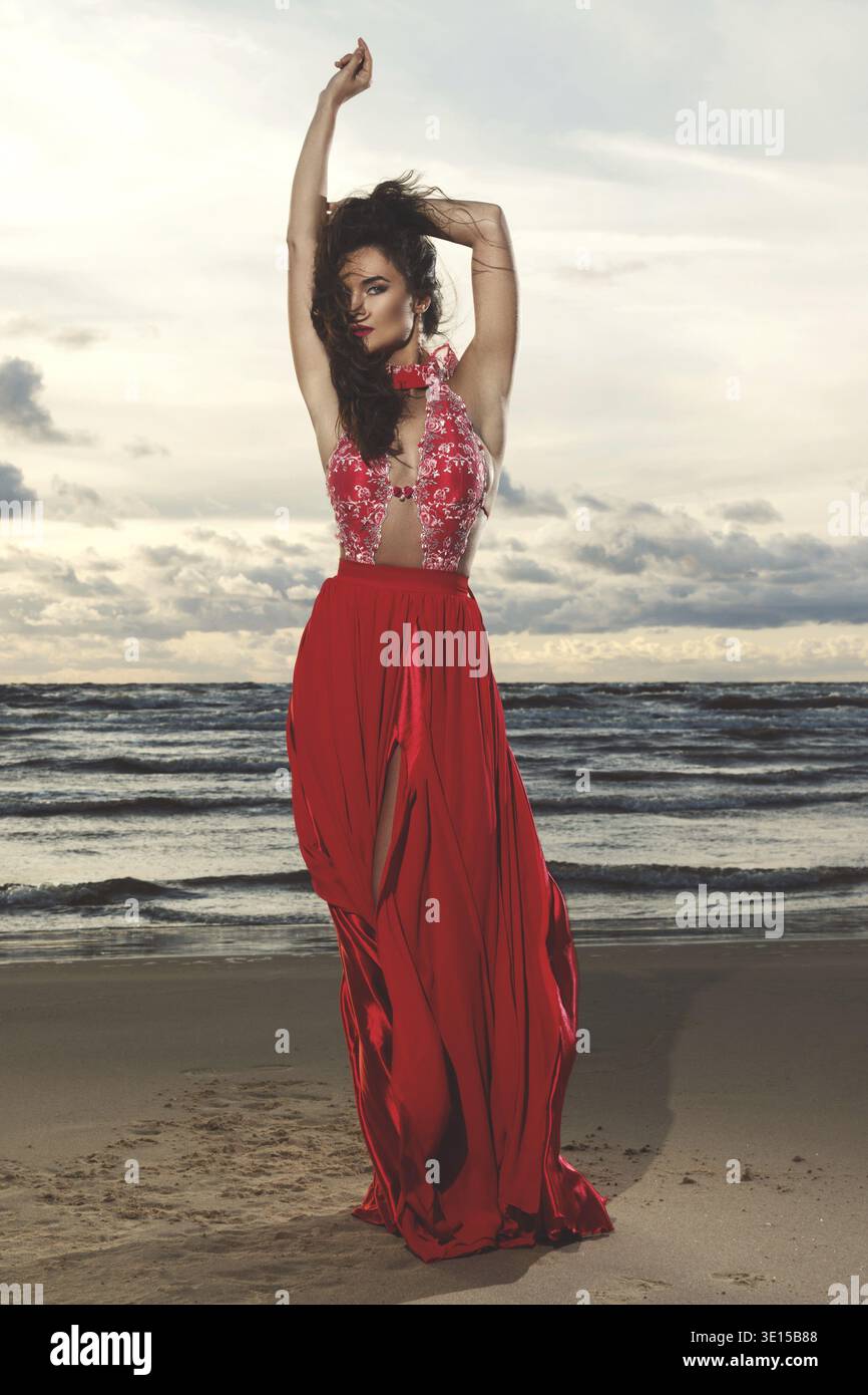 Stunning woman wearing beautiful red dress on the beach during sunset time Stock Photo