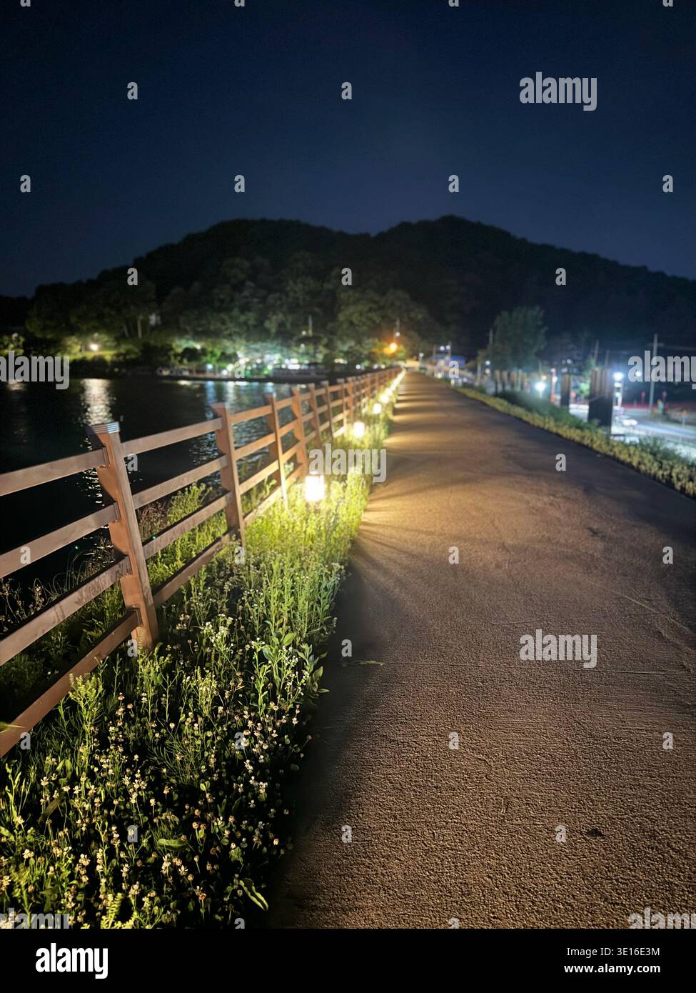 Illuminated wooden pedestrian bridge at night, glowing lanterns along railing, river reflection, Sinjeongho Tourist Park, South Korea - Smartphone Captured Stock Image