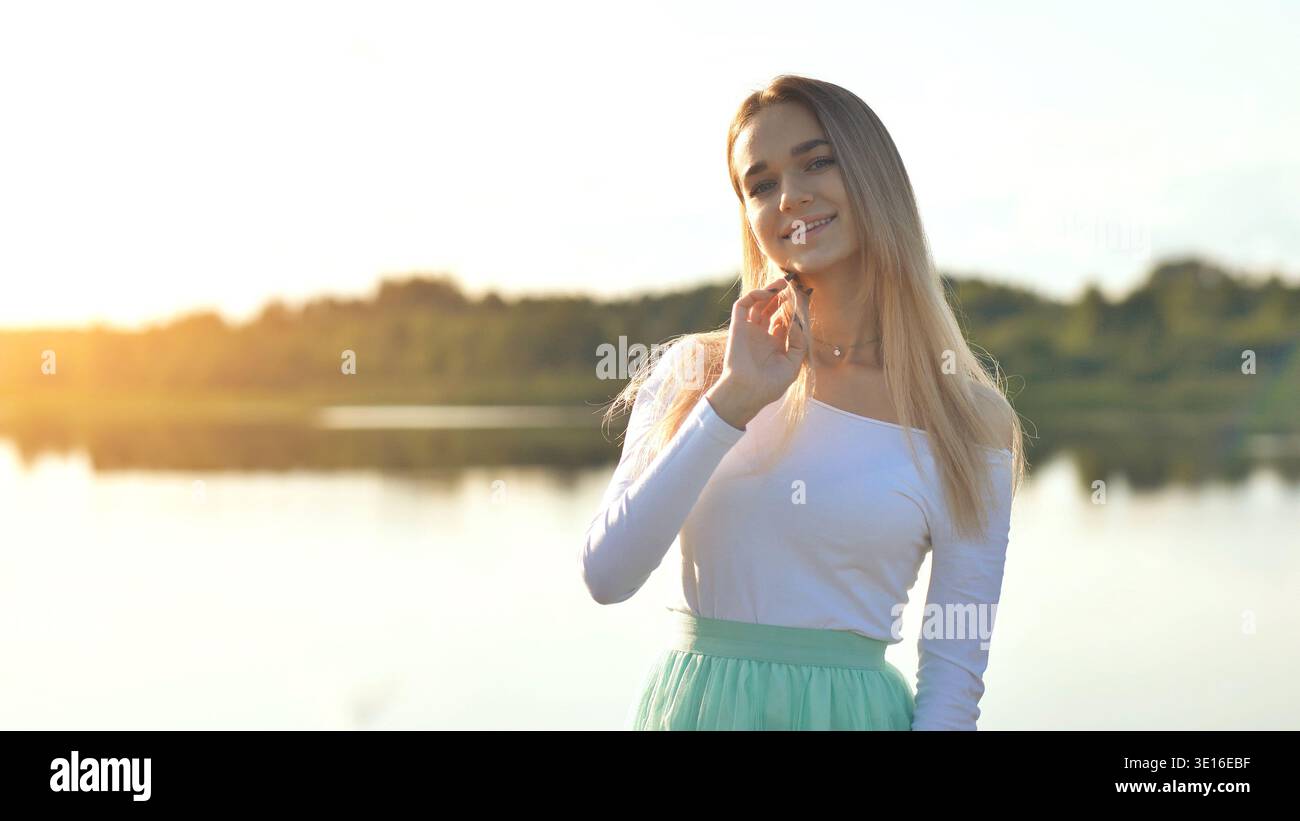 Young woman smiling, enjoying golden sunset hour by peaceful lake Stock Photo
