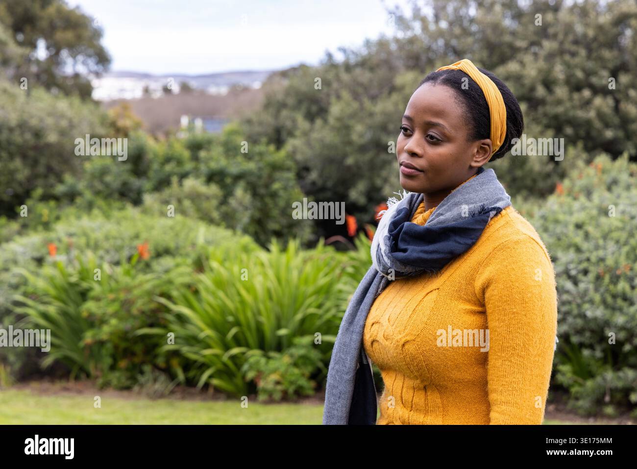 African American woman standing in park wearing mustard-yellow sweater and gray scarf, copy space. Garden, outdoor, greenery, foliage, blossoms, headband, knit || Model released Stock Photo