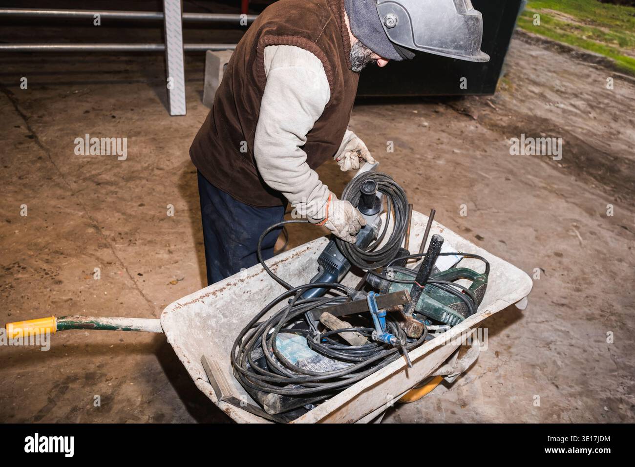 A worker in protective clothing organizes electrical tools and cables in a wheelbarrow at a construction site, preparing for maintenance or repair tas Stock Photo