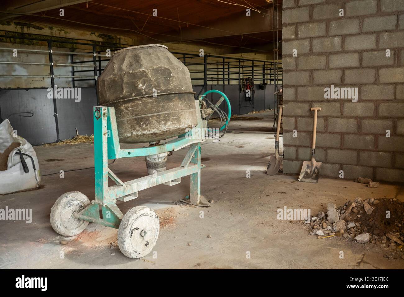 Inside a partially constructed building, a cement mixer and various tools, including shovels, lie on a dusty floor, showcasing ongoing construction an Stock Photo