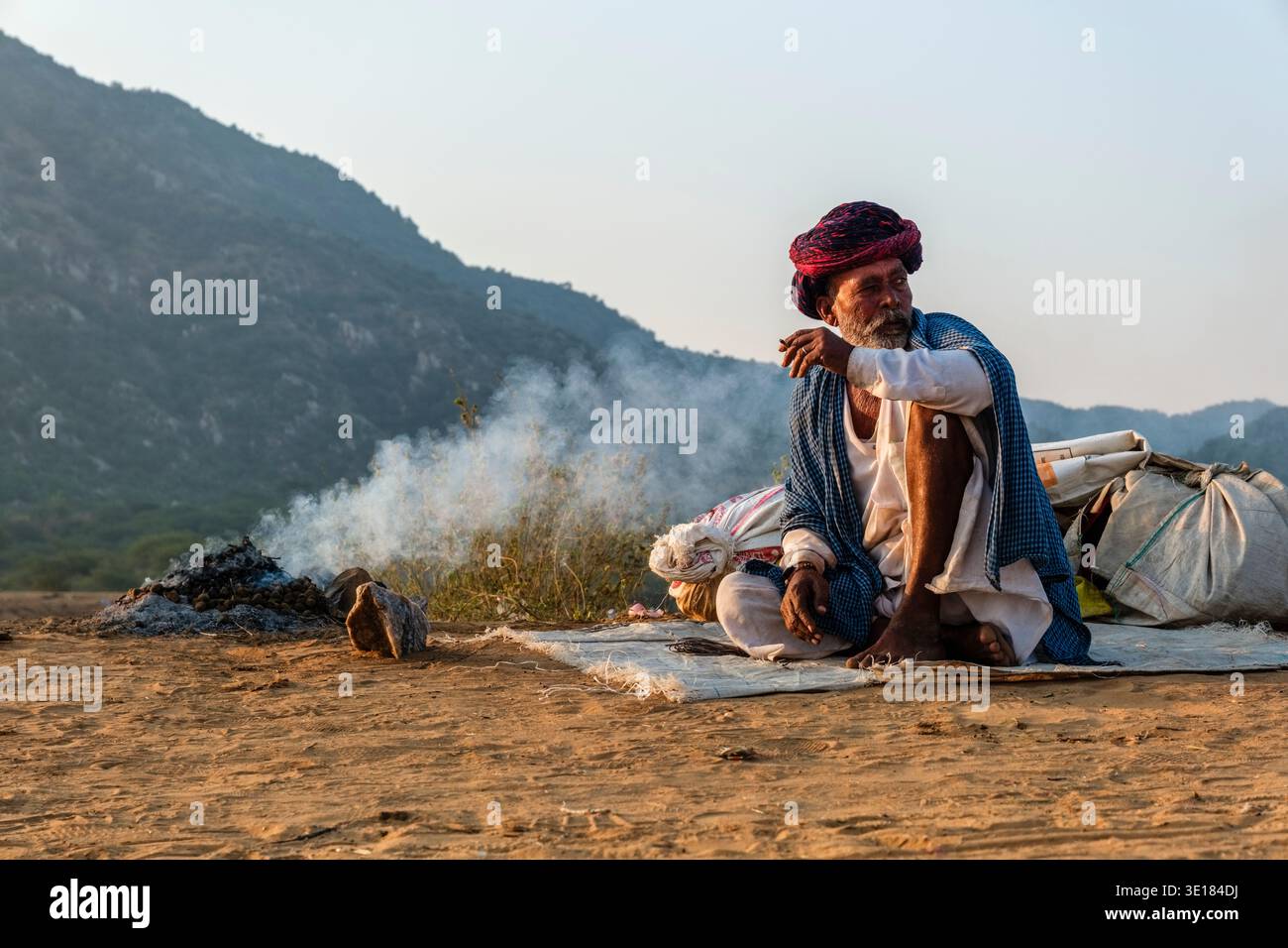 A man sits on a mat next to a small smoking fire at the Pushkar Camel Fair with the silhouette of a large mountain visible in the distance. Stock Photo