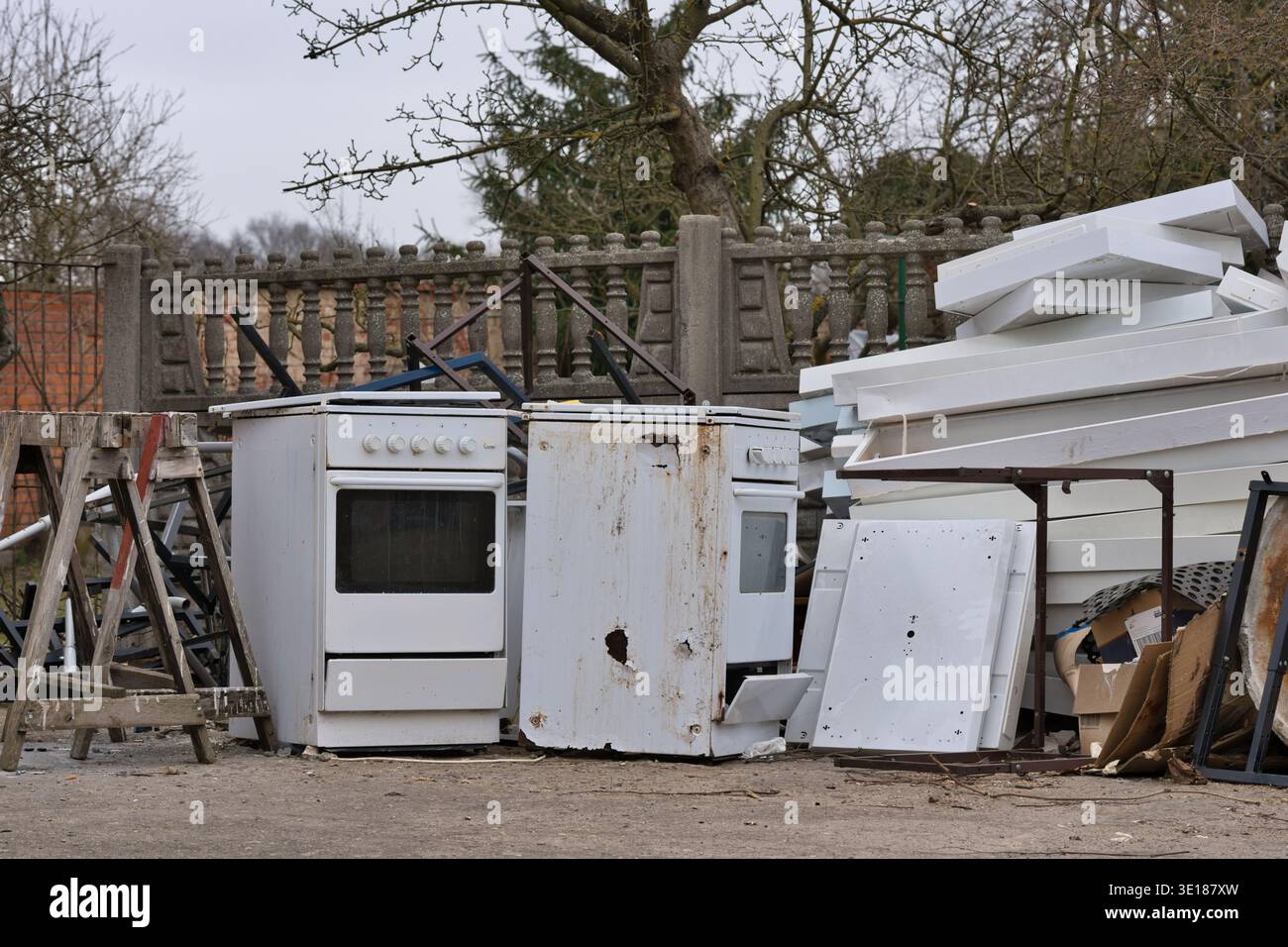 Old, rusty gas stoves and white bulky waste stored outdoors near a concrete fence. Electronic waste and scrap metal Stock Photo