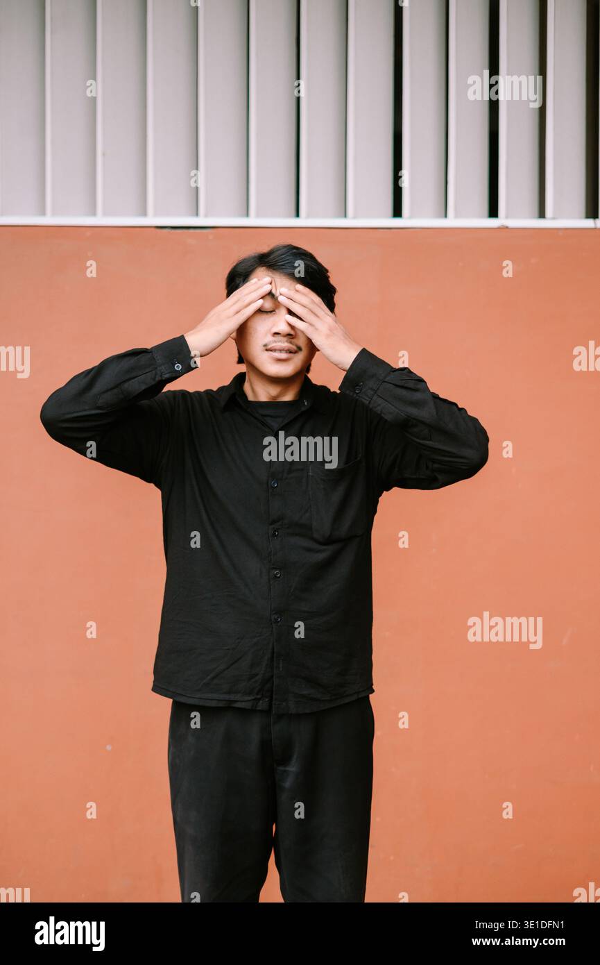 A young man covers his eyes with his hands, displaying feelings of stress, fatigue, or discomfort. He stands against a vibrant orange wall. Stock Photo