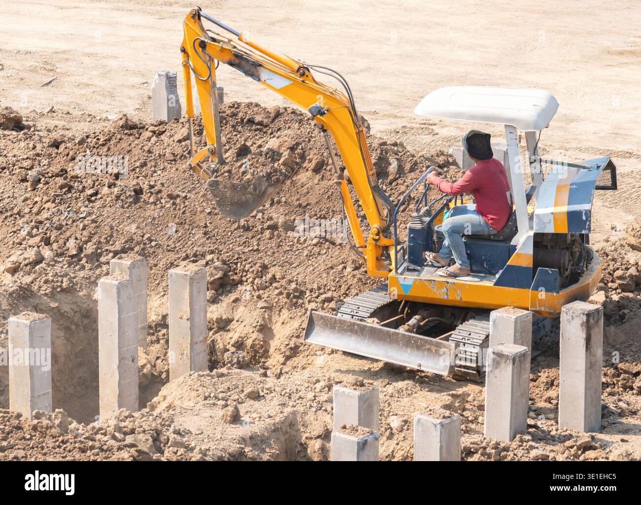 Small yellow excavator digging soil around concrete foundation piles at ...