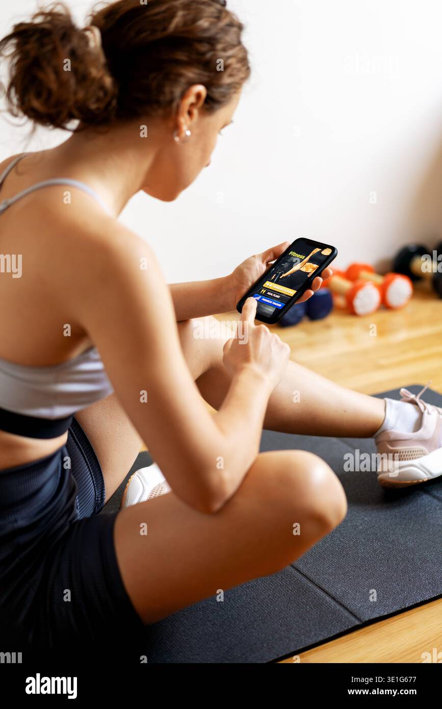 Fit woman sitting on yoga mat and launching mobile fitness app on her smartphone before indoor workout Stock Photo