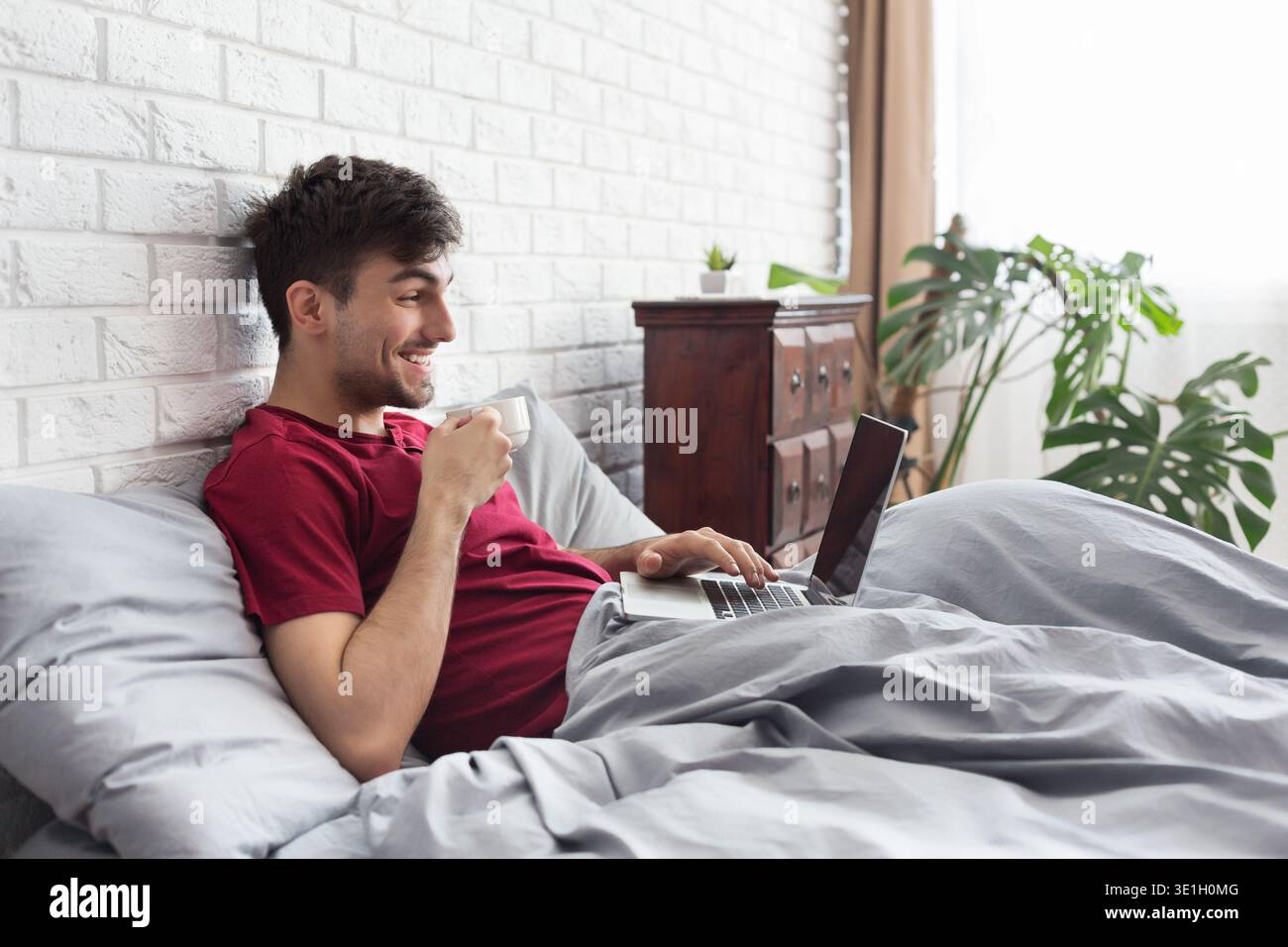 Happy man drinking coffee and using laptop in bed Stock Photo