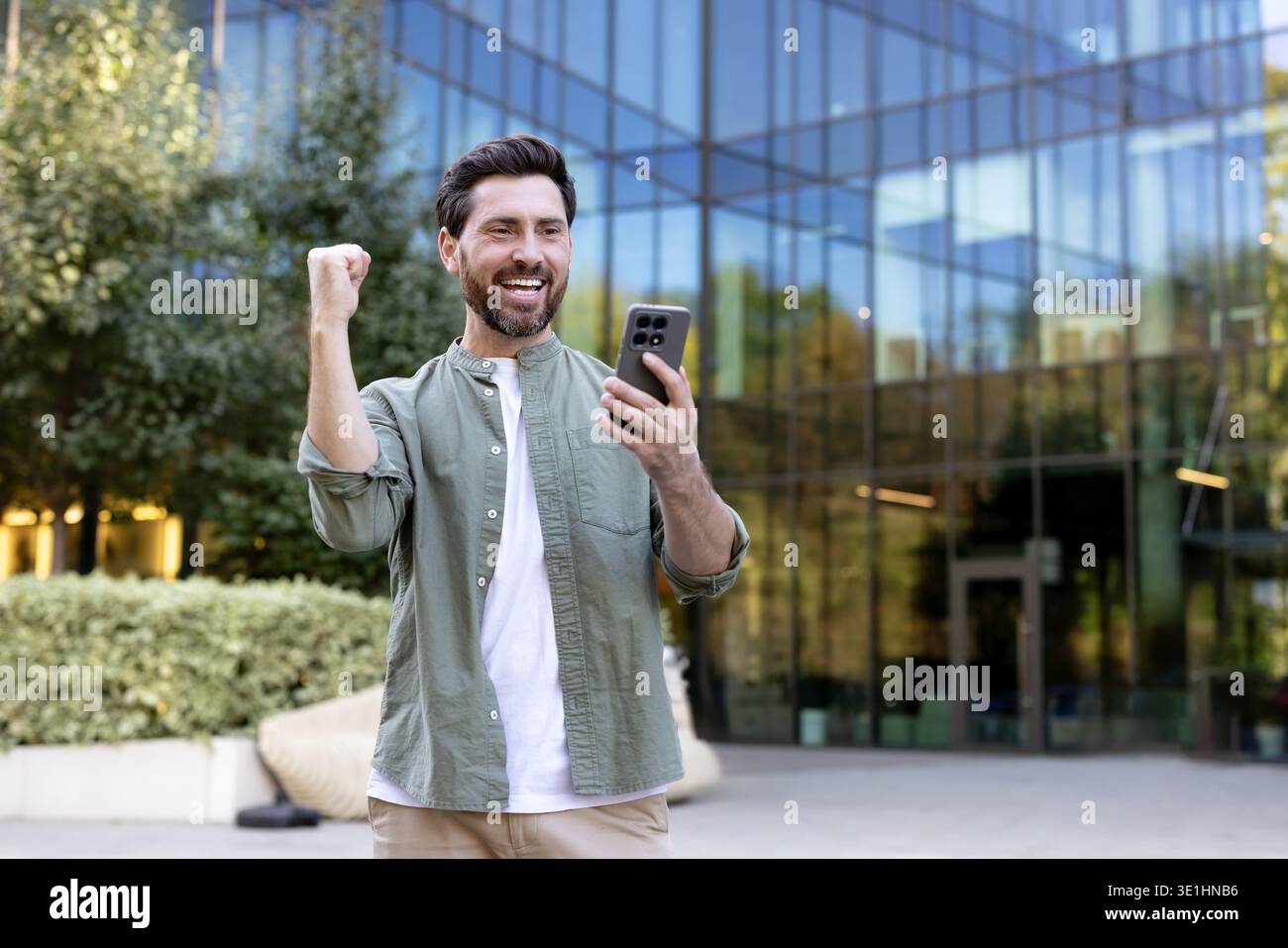 Excited businessman cheering and raising his fist while looking at a smartphone screen, standing outside a modern office building, celebrating a professional victory or good news Stock Photo