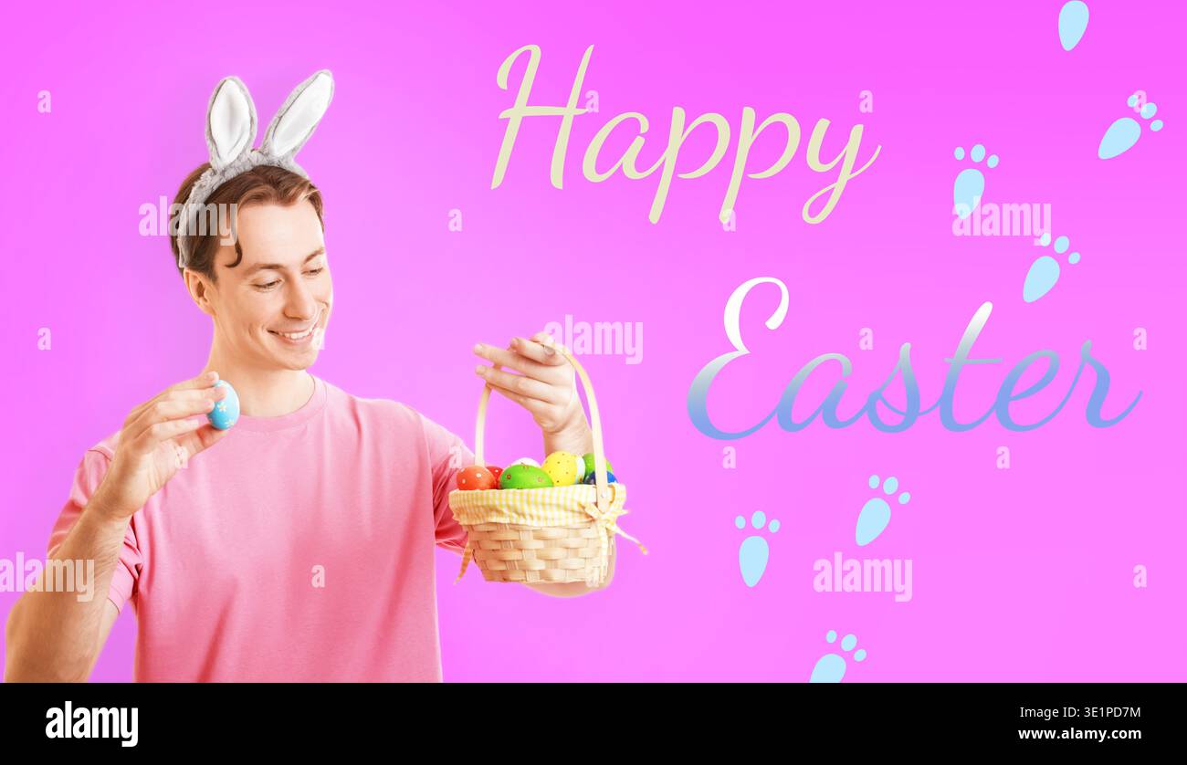 Cheerful man in bunny ears holding a blue Easter egg and a basket full of colorful eggs against a pink background with Happy Easter text and paw print Stock Photo