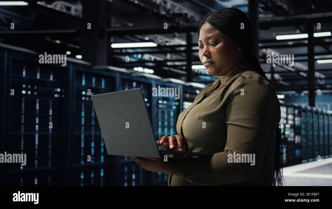 Software developer in data center using laptop to upgrade systems for increased stability. Server hub employee configuring equipment rigs with notebook for optimum performance, camera A Stock Photo