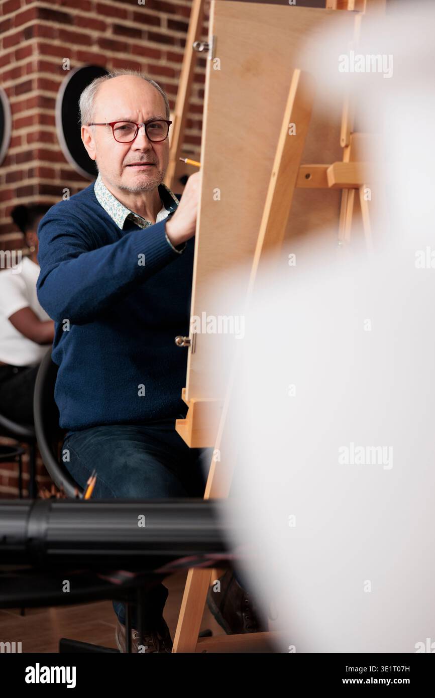 Concentrated focused senior man sitting at easel drawing object with graphite pencil. Mature student looking at ceramic vase making sketch on canvas during group art class in studio Stock Photo