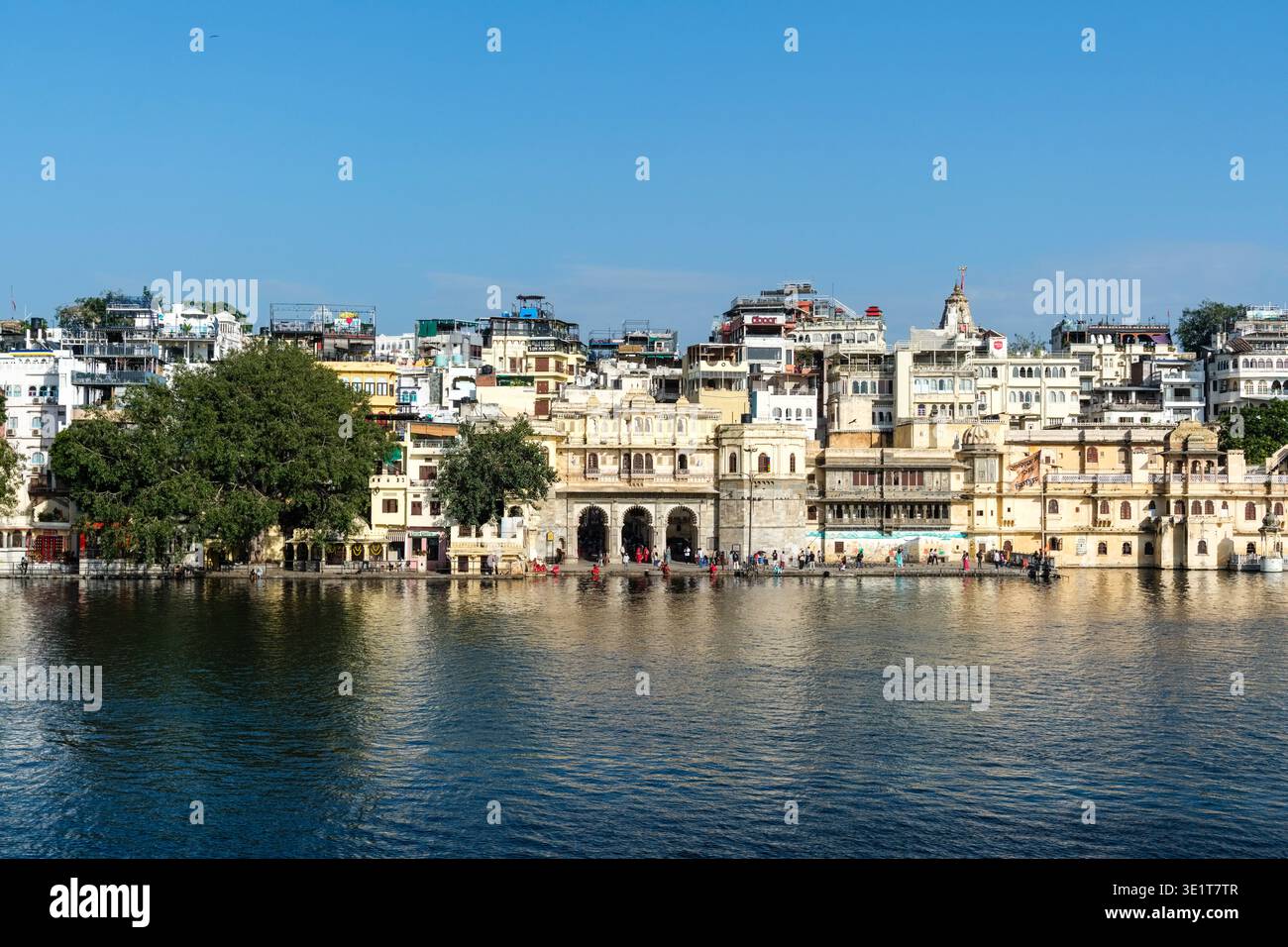 People gather at Gangaur Ghat in Udaipur, where historic stone buildings and temples line the waterfront of Lake Pichola under a clear blue sky. Stock Photo