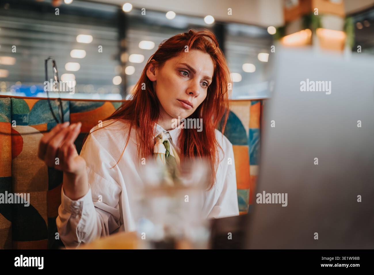 Business worker redhead looking at laptop in a cafe while working remotely and holding glasses Stock Photo