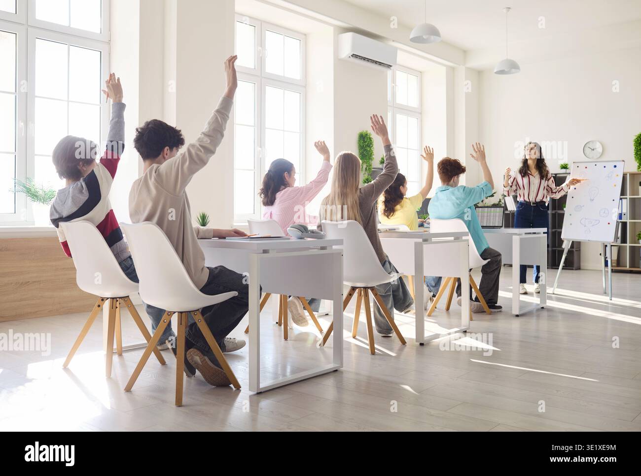 High School Students Raise Hands in Bright Classroom while Teacher Guides Class Stock Photo