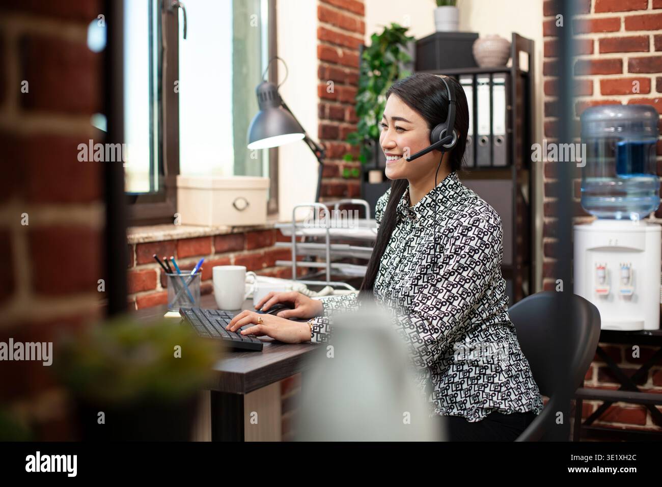 Female employee uses desktop pc, typing on keyboard and engaging in a lively conversation through wired headphones in modern workplace. Smiling woman seated at office desk, using computer and headset. Stock Photo