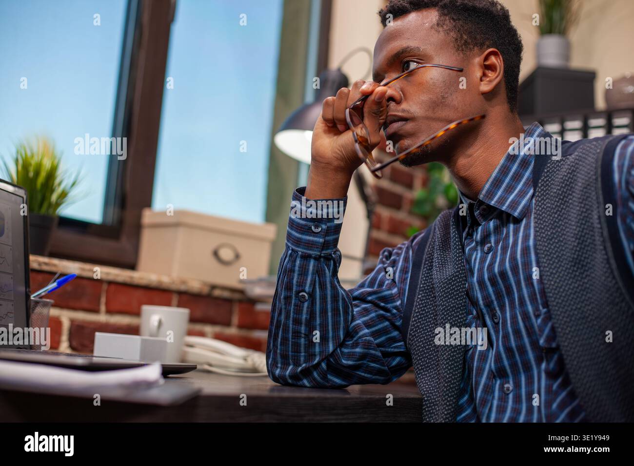 Overwhelmed male analyst holding his eyeglasses and looking outside the window at startup company workspace. Businessman with spectacles in hand, sitting at desk, deeply thinking about strategy ideas. Stock Photo