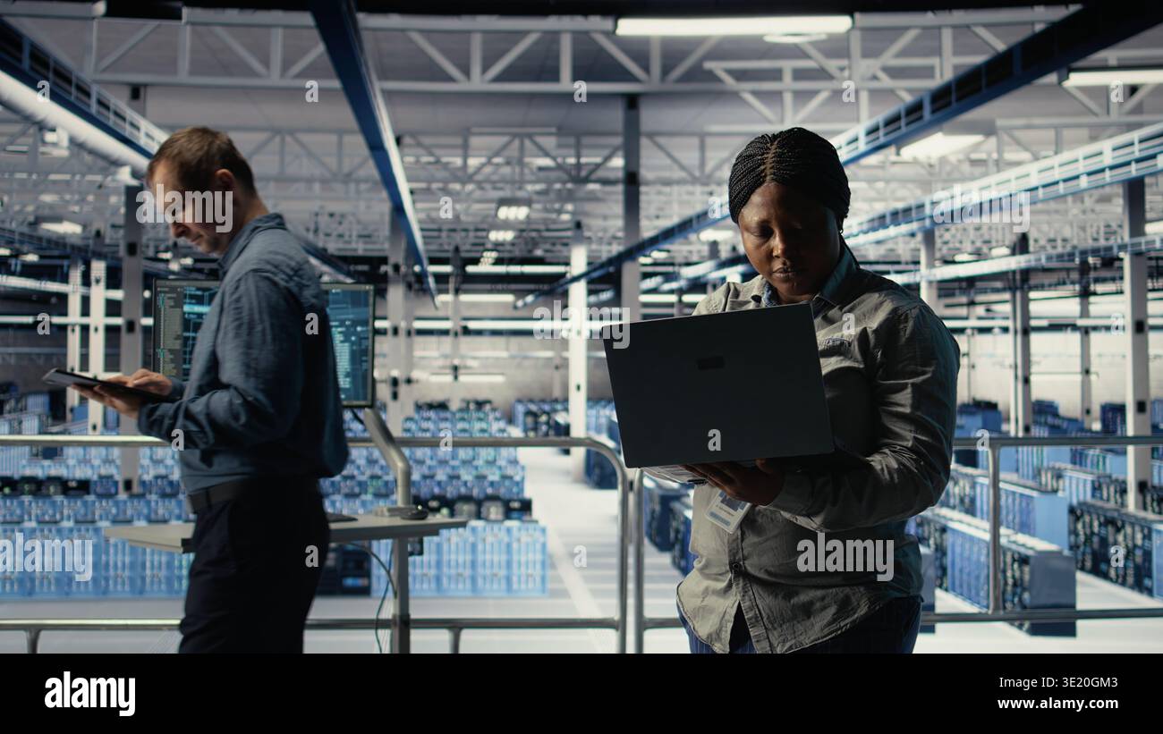 Data center technician deploys machine learning software on infrastructure racks. African american woman in server farm trains AI systems with datasets using laptop to support applications, camera A Stock Photo