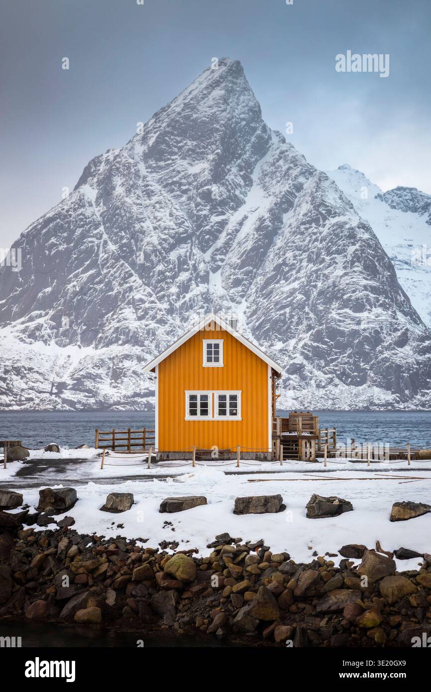 View of the Famous Yellow House with view of Olstind peak covered with snow in the background. Sakrisoy, Reine, Nordland, Lofoten Islands, Norway. Stock Photo