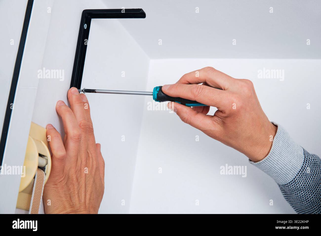 a man tightens a screw with a screwdriver to secure a black metal bracket on a white wall, holding the bracket steady with his other hand Stock Photo