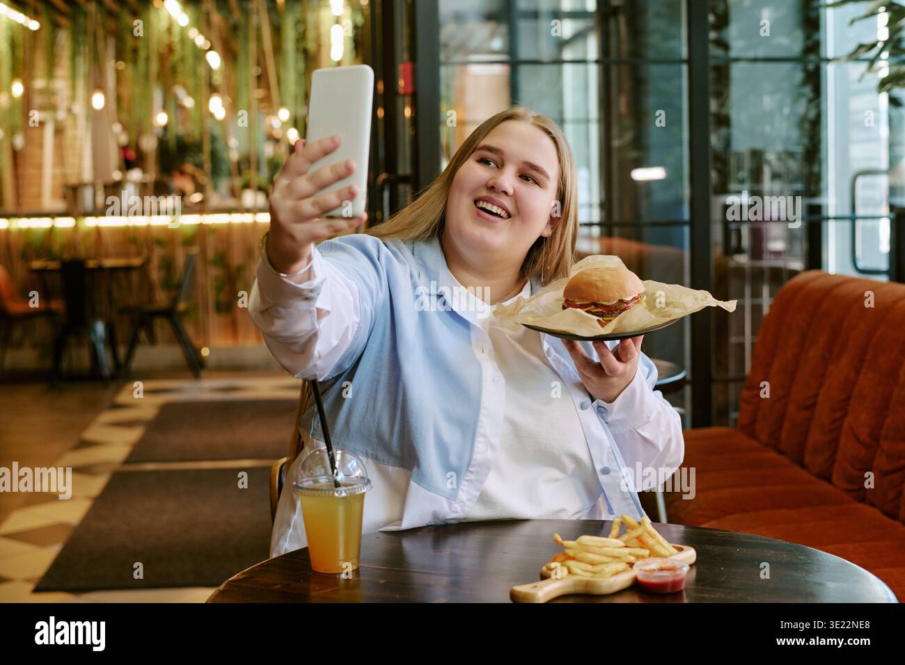Young Woman Taking Selfie While Holding Burger in Restaurant Stock Photo
