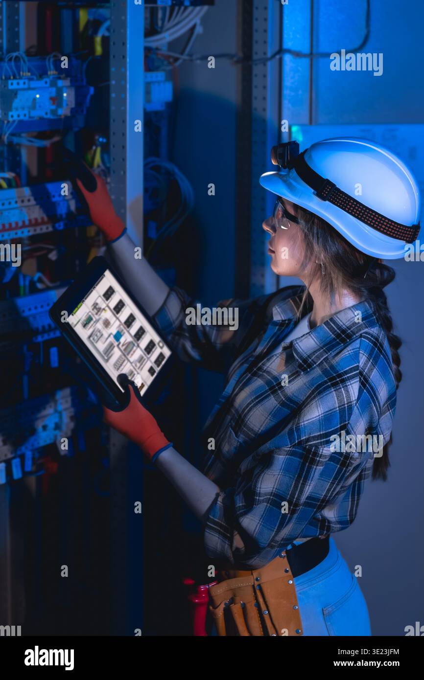 Female electrician inspecting electrical panel with tablet in server room. Power engineer. Stock Photo