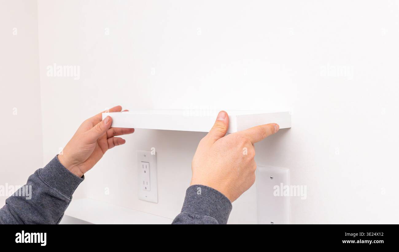Mans hands holding white rectangular shelf plank against white wall near installed black bracket. Home improvement, positioning shelf board, DIY Stock Photo
