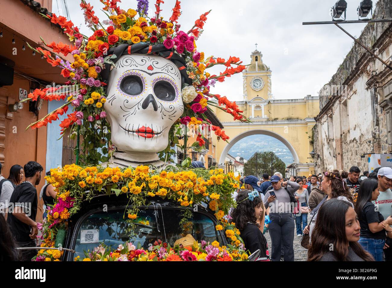 Festival De Flores - Antigua Guatemala - A large decorated sugar skull float covered in vibrant flowers leads a crowded street parade under the iconic Stock Photo