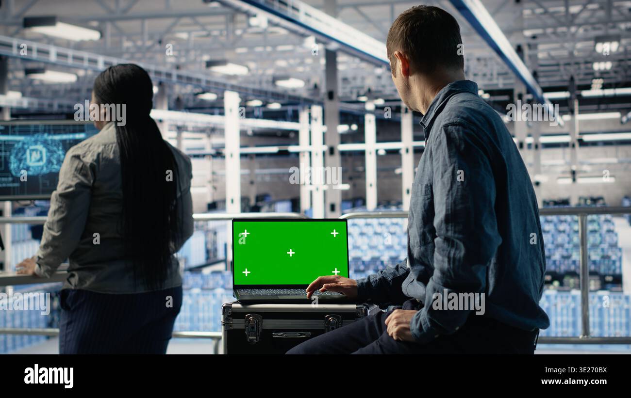 Data center IT experts investigate AI configurations using green screen laptop, making adjustments. Server hub engineers check deep learning errors on chroma key notebook, camera B Stock Photo