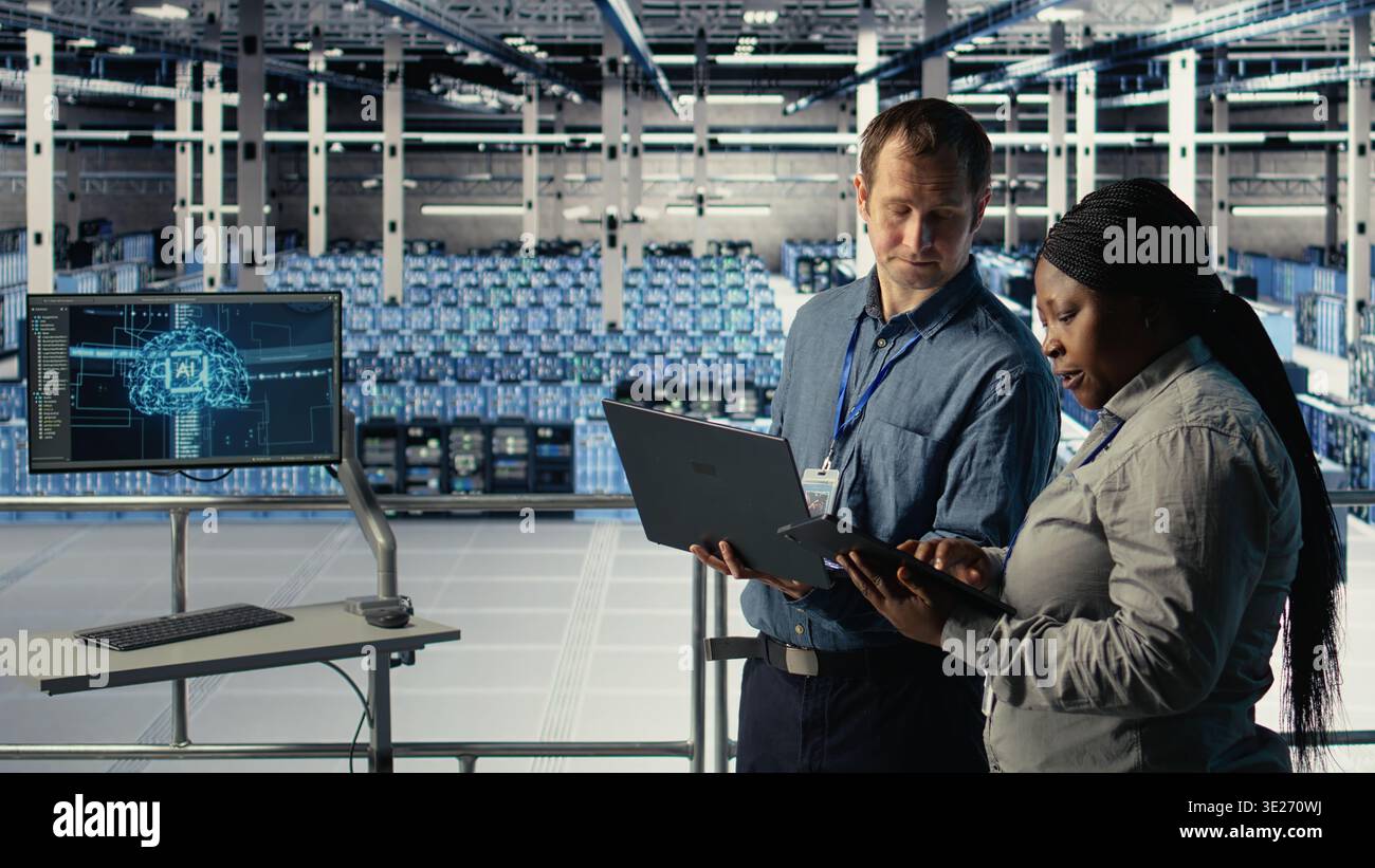 Server room coworkers use laptop to install updates and keep AI systems running. Data center engineering team uses notebook to write code improving infrastructure output, camera A Stock Photo
