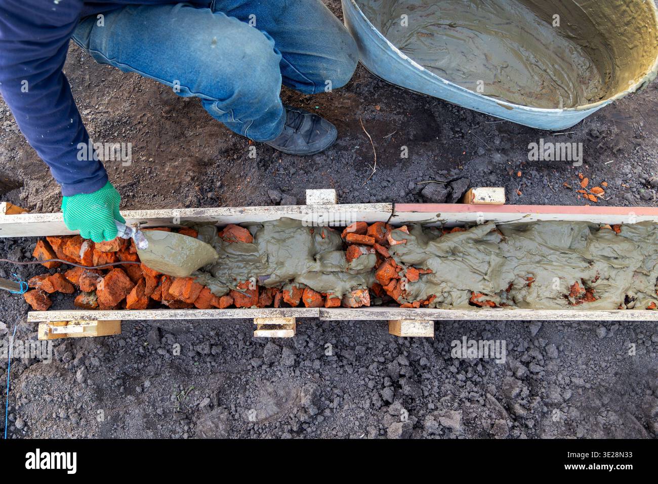 Pouring concrete into brick foundation form. Top view of a worker spreading wet concrete over broken bricks inside wooden formwork during small Stock Photo